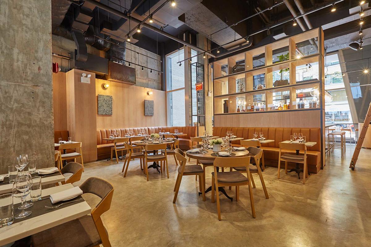 Modern restaurant interior with wooden tables and chairs, tan cushioned booths, large windows, decorative shelves with plants and bottles, and ambient lighting from ceiling fixtures. Tables are set for dining.