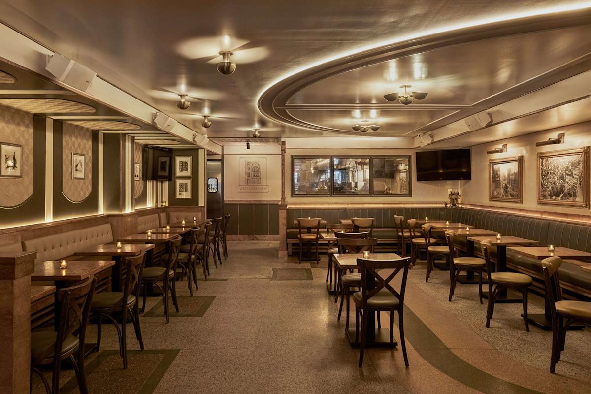 A warmly lit, empty restaurant with wooden tables and chairs, green upholstered walls, framed art, and curved ceiling lights, creating a cozy and inviting atmosphere.