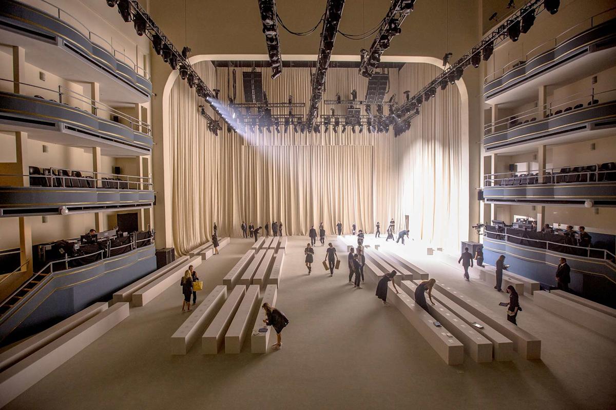 People are setting up and walking around a large, elegant theater with tall curtains, high ceilings, and tiered balconies. White benches are arranged on the floor, illuminated by soft lighting.