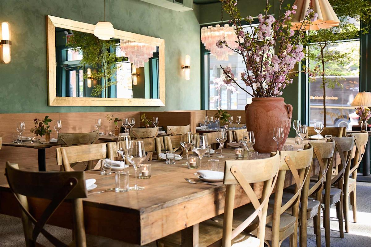 A cozy restaurant interior with wooden tables and chairs, set with glassware and napkins. Large vase with pink flowers is on the main table. A mirror and hanging lights decorate the green-painted walls. Natural light fills the room.