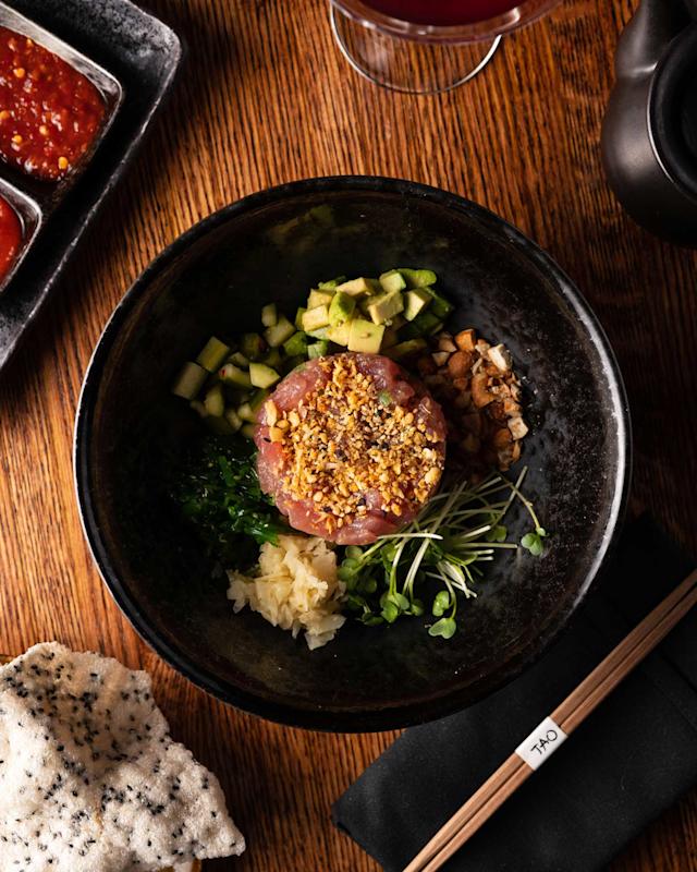 A black bowl filled with diced raw fish topped with crunchy bits, surrounded by chopped cucumber, avocado, sprouts, pickled ginger, and greens, set on a wooden table with chopsticks, a crisp chip, and dipping sauces nearby.