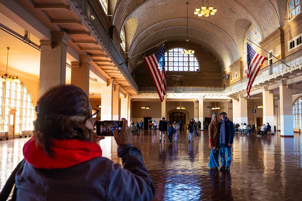 A person in a red hoodie takes a photo of two people posing inside a large, sunlit hall with arched windows, high ceilings, and two large American flags hanging from the ceiling. Other visitors are seen in the background.