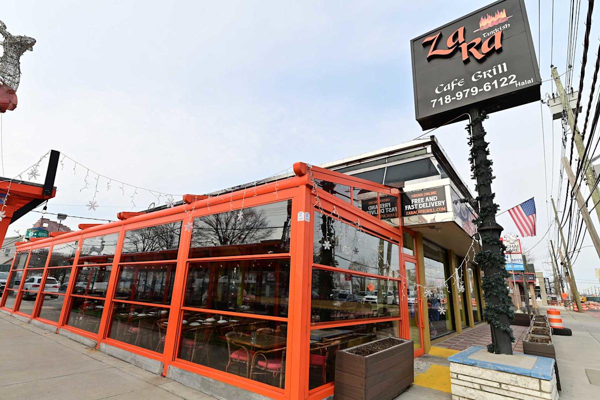 A street view of LaRra Cafe Grill with a black sign, orange-framed outdoor seating area, glass windows, and American flag nearby on a cloudy day.