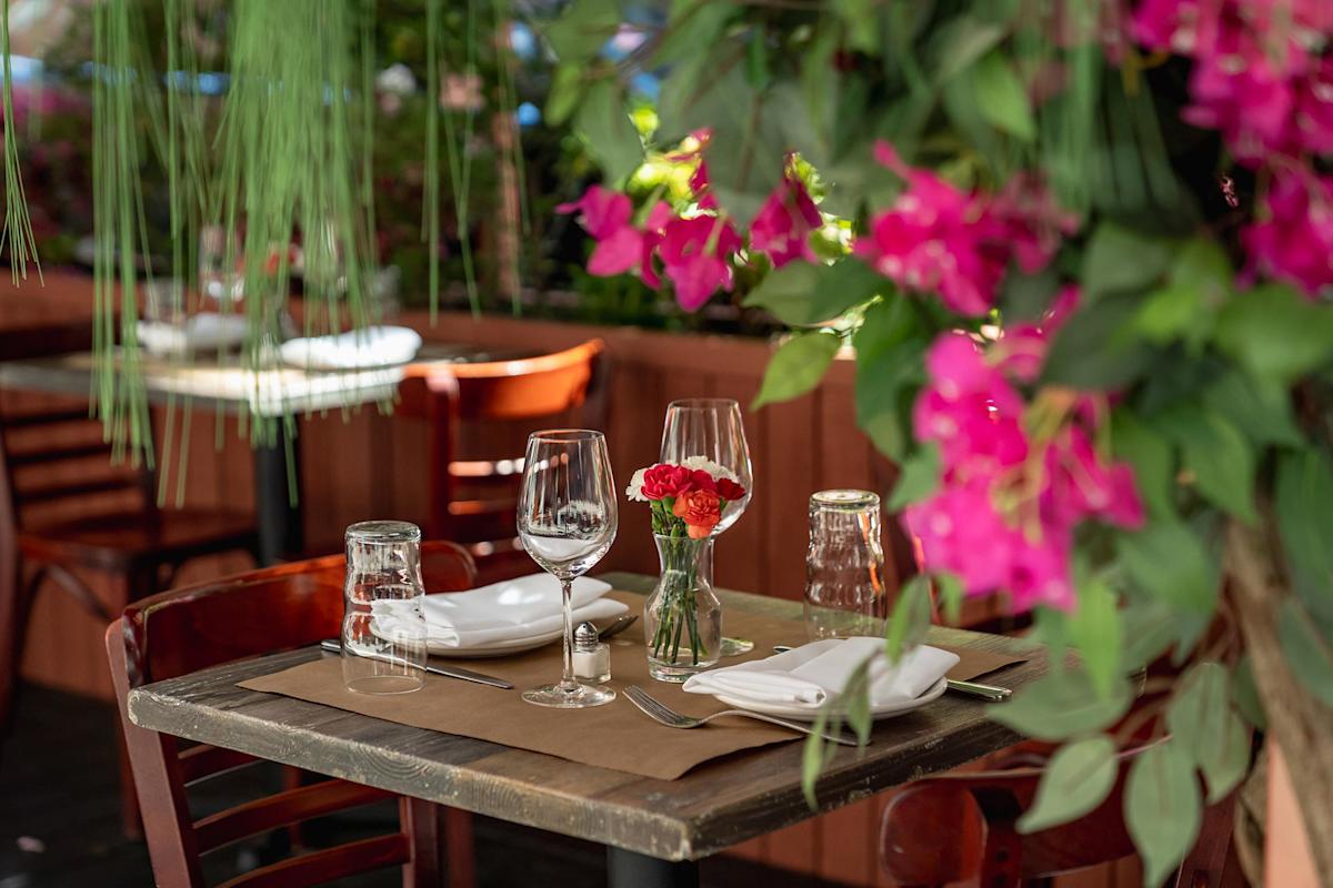 A table set for two with wine glasses, plates, and a small vase of red flowers, surrounded by lush pink and green foliage in an outdoor restaurant setting.