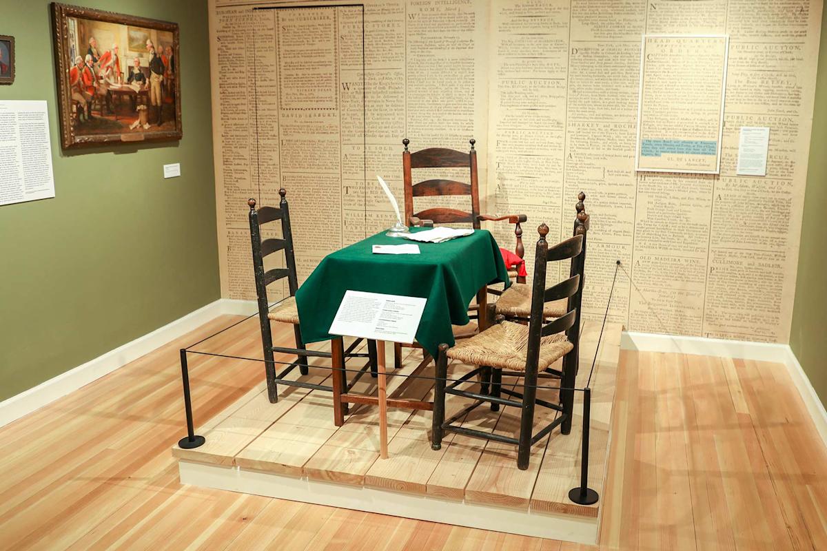 A museum exhibit featuring a table with a green cloth, three wooden chairs, historical documents, and a quill pen, set against walls covered with handwritten text. A framed painting hangs on the green wall.