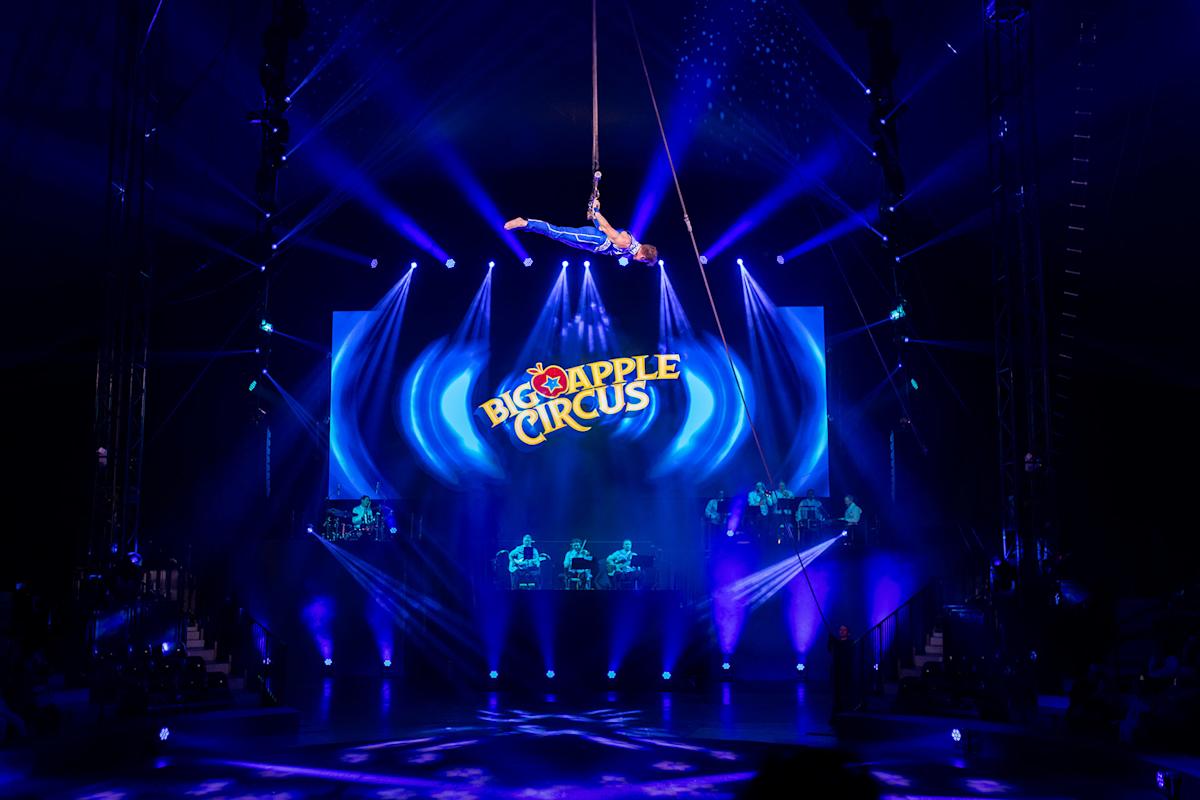 An aerial performer hangs from a rope high above a brightly lit stage at the Big Apple Circus, with blue spotlights, a live band below, and the circus logo illuminated in the background.