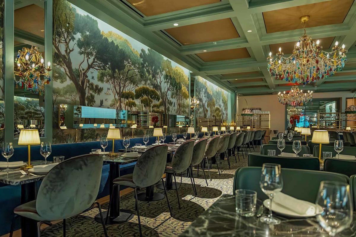 Elegant restaurant interior with chandeliers, velvet chairs, marble tables set for dining, and a mural of trees along the wall, creating a luxurious and inviting atmosphere.