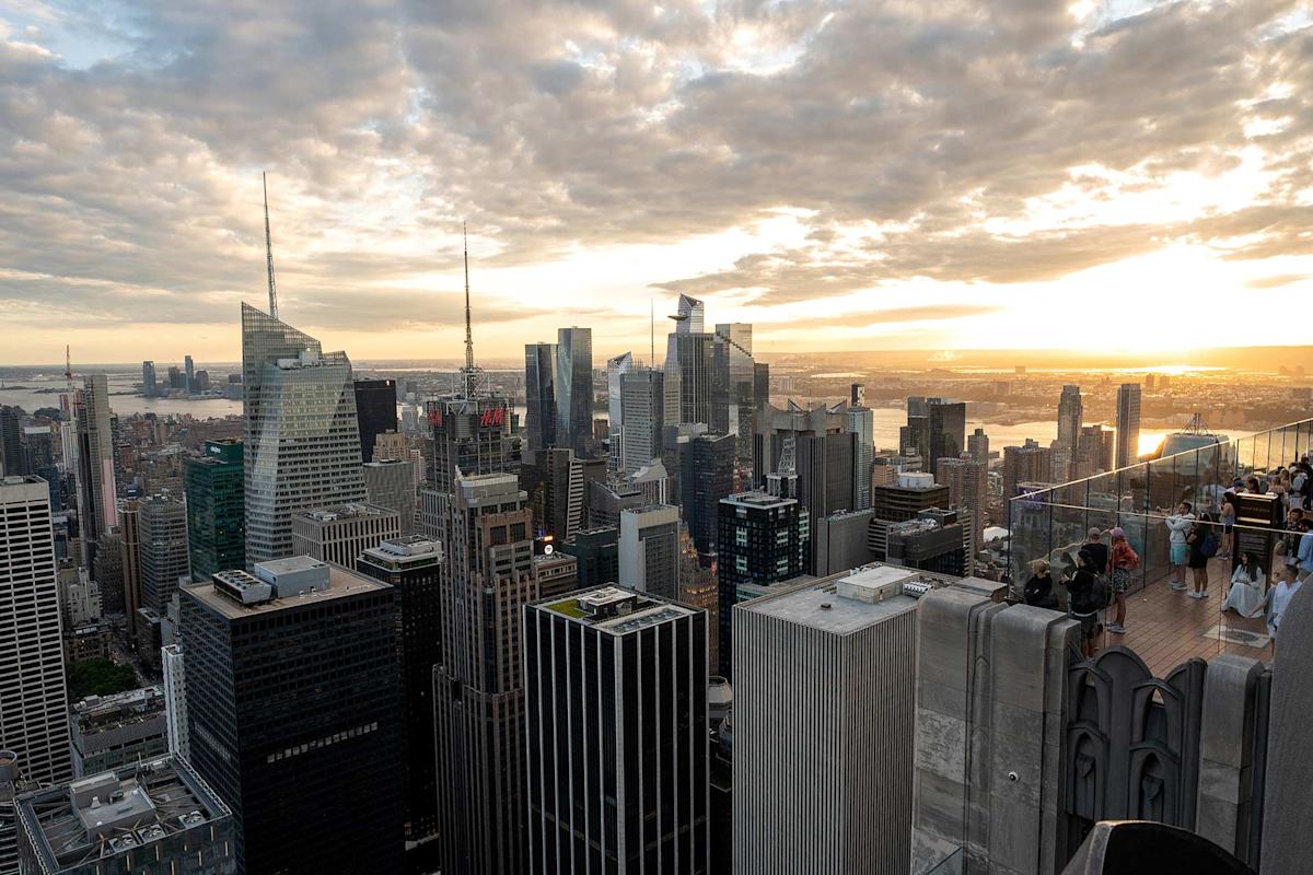 A view of the New York City skyline at sunset, with tall skyscrapers under a partly cloudy sky and people standing on an observation deck in the foreground.