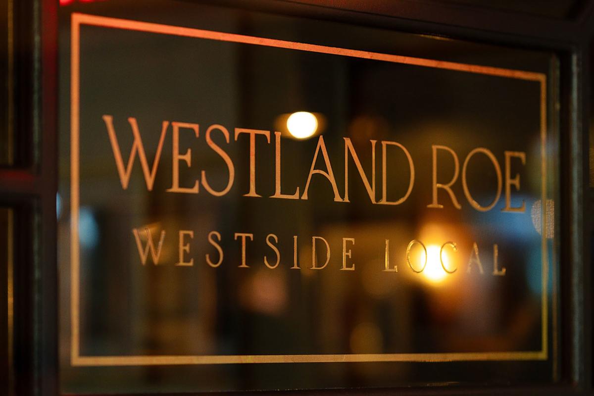 A glass window sign with gold lettering reads "WESTLAND ROE WESTSIDE LOCAL" against a dark blurred background with warm lighting.