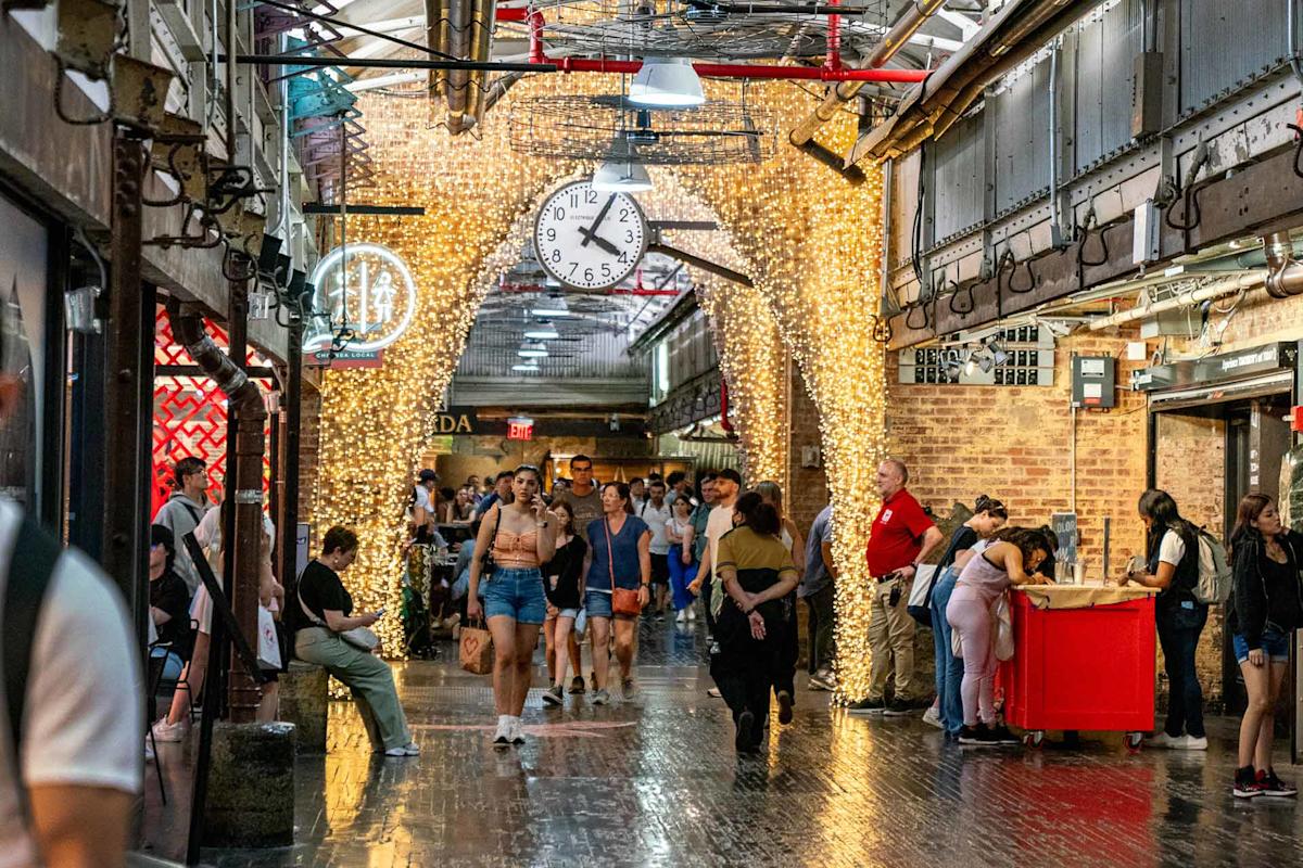 A busy indoor market with people walking and shopping, illuminated by string lights wrapped around brick archways. A large clock hangs from the ceiling, and various stalls and shops line the corridor.