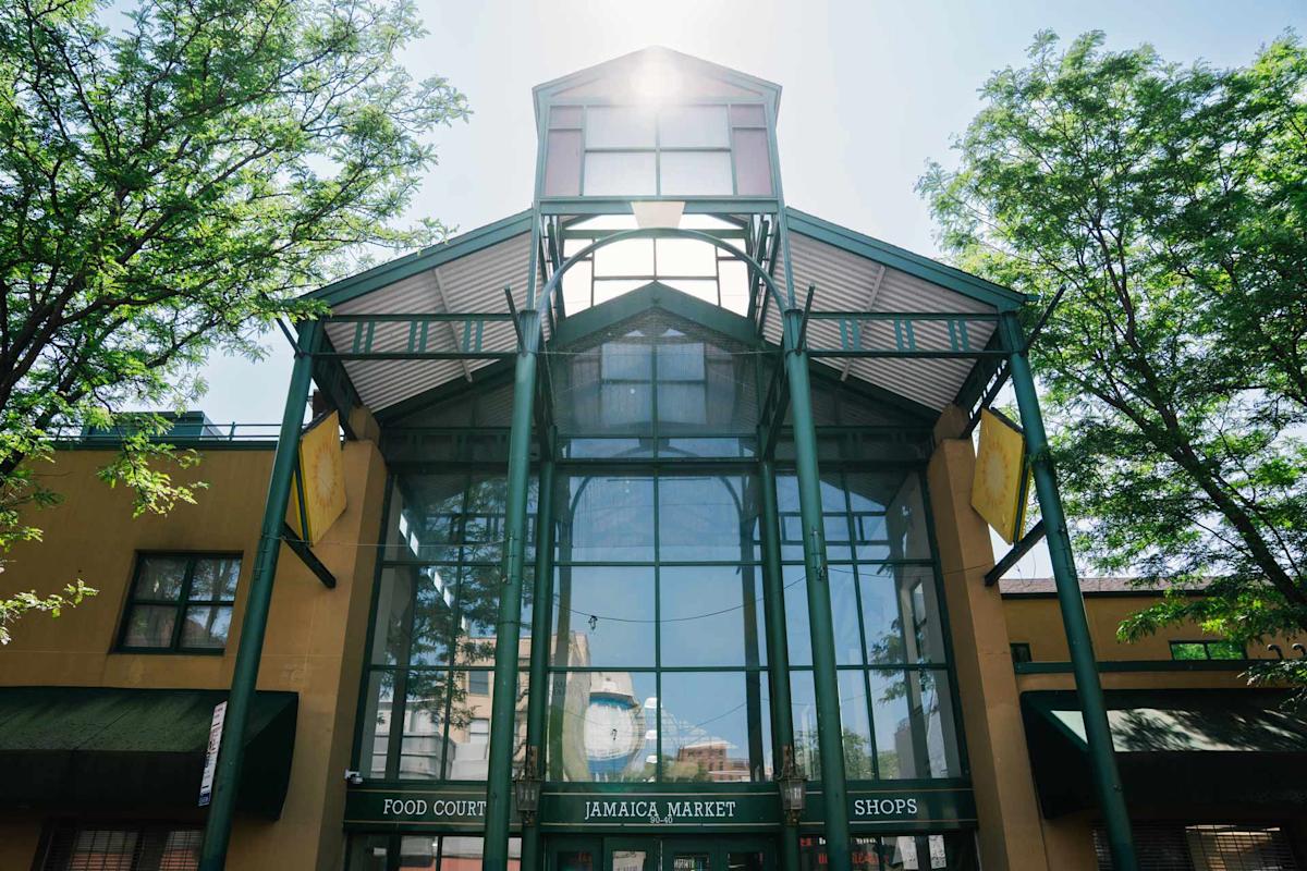 Glass entrance of Jamaica Market with green metal framework, trees on both sides, and sunlight shining from above. Signs read "Food Court," "Jamaica Market," and "Shops.