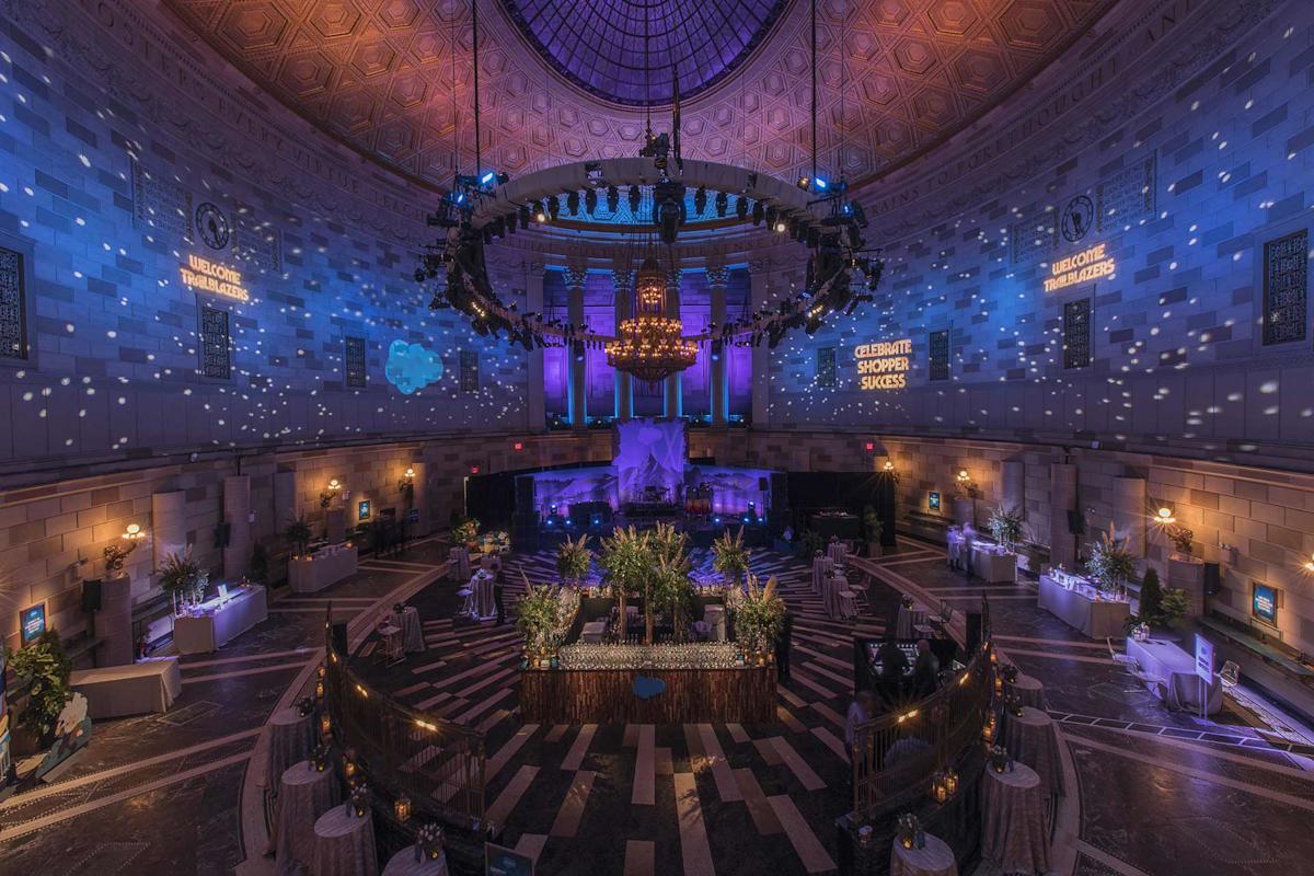 A grand, circular event hall with vaulted ceilings, blue and purple lighting, a large chandelier, scattered tables, floral centerpieces, and decorative spotlights creating a festive atmosphere.