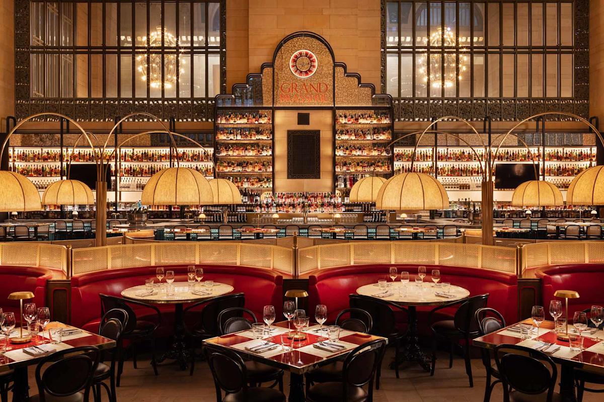 Elegant restaurant interior with red leather booths, round tables set with glasses and cutlery, art deco lighting, and a grand bar with illuminated shelves lined with bottles. Large windows and chandeliers add to the upscale ambiance.