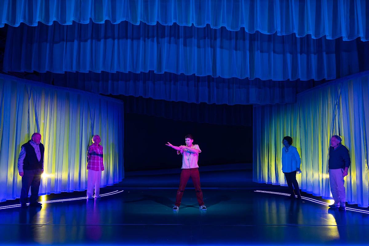 A person in a pink shirt stands center stage under blue and purple lights, gesturing, while four others stand apart in the background near softly lit, wavy curtains. The scene feels dramatic and theatrical.