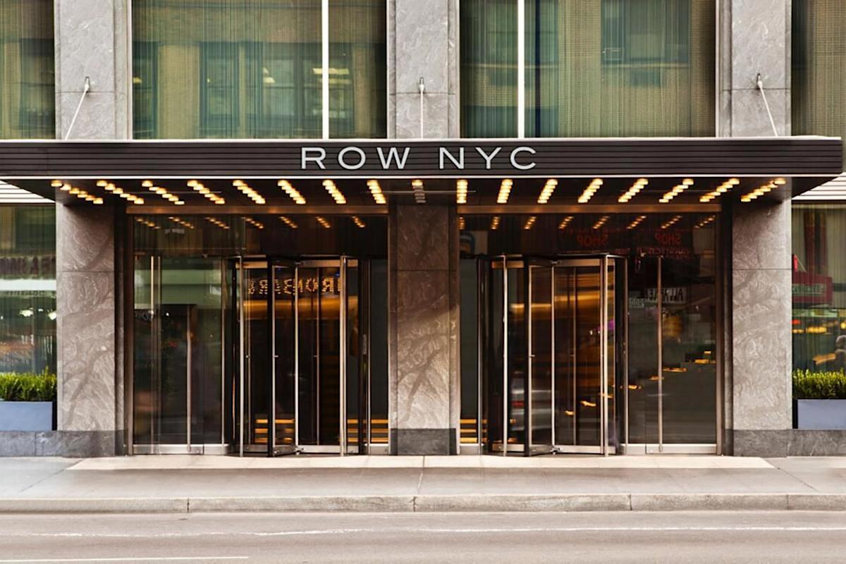 Entrance of the Row NYC hotel with a modern glass facade, metal accents, and a canopy with lights above the revolving doors. The hotel name “ROW NYC” is displayed prominently above the entrance.