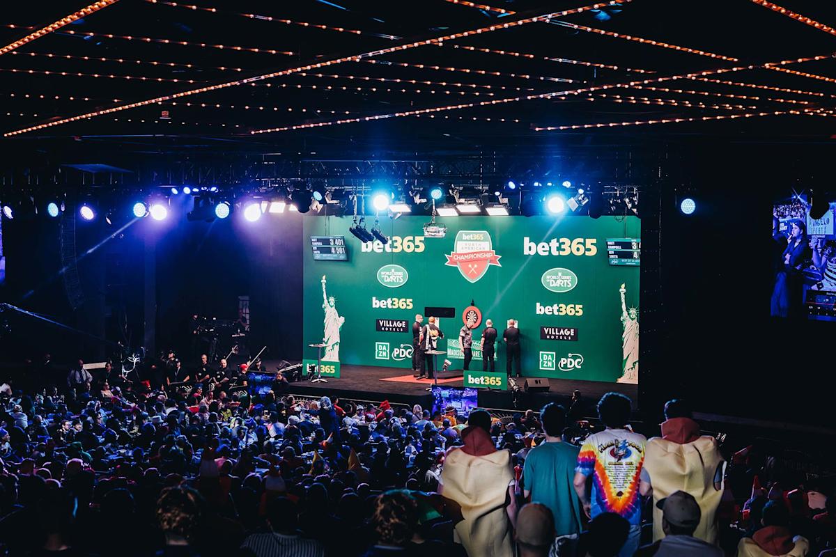 A large crowd watches a live darts event on stage, with bright lighting and a green backdrop displaying sponsor logos. Several people stand on stage, and the audience fills the venue, some in colorful costumes.