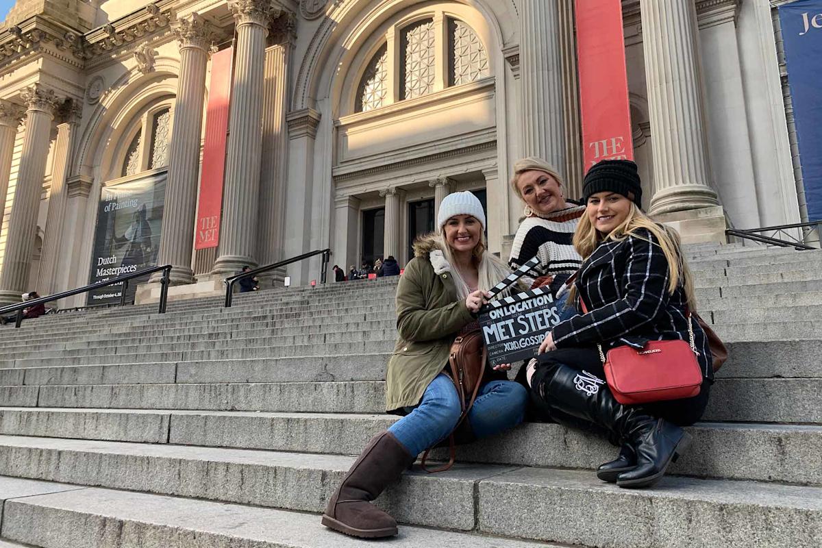 Three women sit smiling on the steps outside The Metropolitan Museum of Art in New York City, holding a sign that says "On Location Met Steps." The museum’s grand columns and banners are visible in the background.