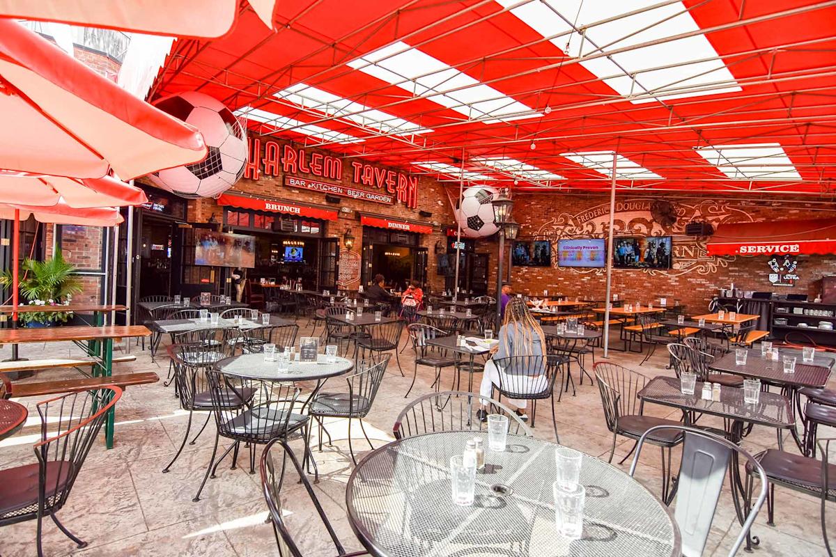 Outdoor seating area at Harlem Tavern, featuring metal tables and chairs under a red canopy, large soccer ball decorations, and brick walls with TVs and signage in the background.
