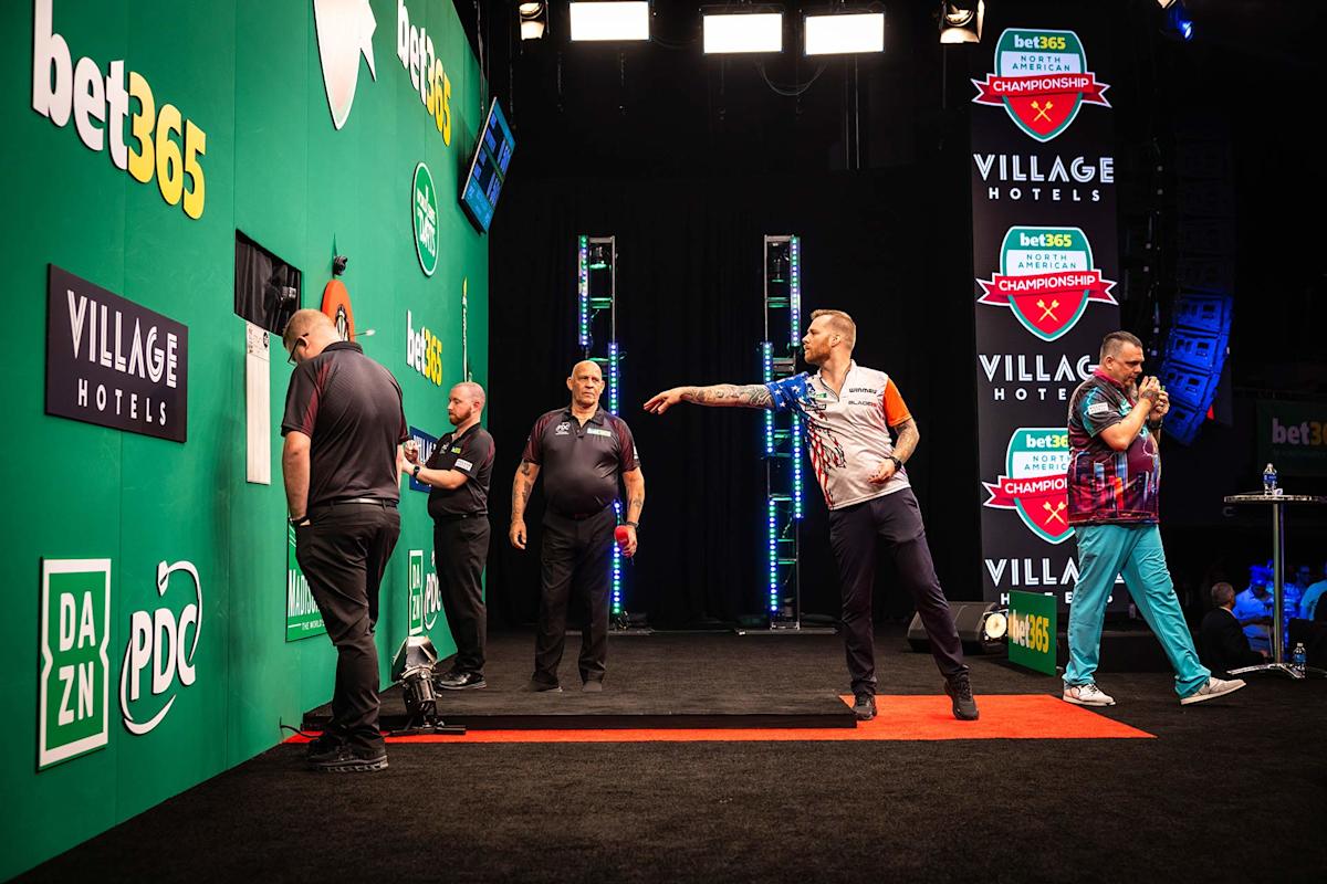 A professional darts match in progress, with one player throwing a dart, others watching, and a referee observing, set on a stage with bet365 and Village Hotels branding.