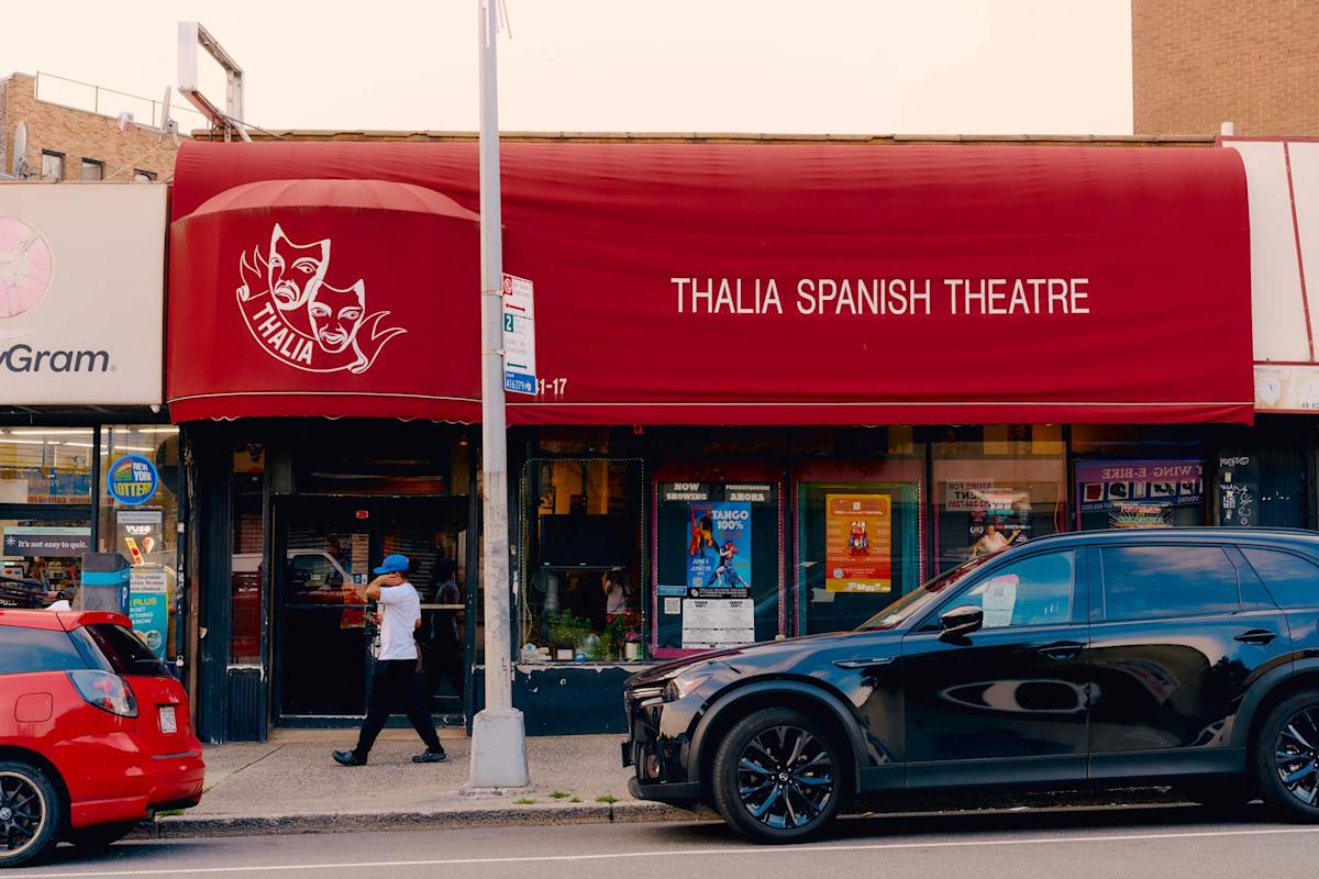 Street view of Thalia Spanish Theatre, with a red awning displaying its name and logo. A person walks by the entrance, and two cars are parked along the curb in front of the building.