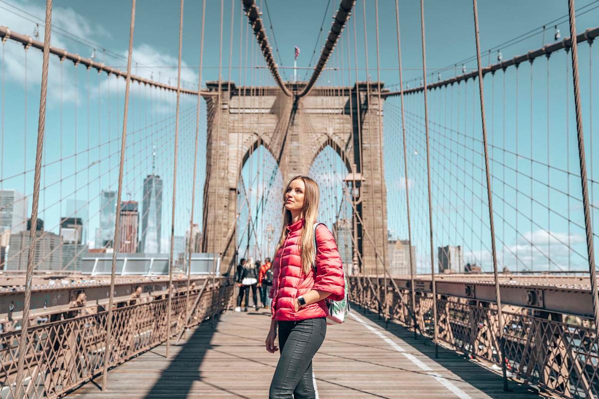 A woman in a red jacket stands on the Brooklyn Bridge with New York City skyscrapers in the background under a clear blue sky.