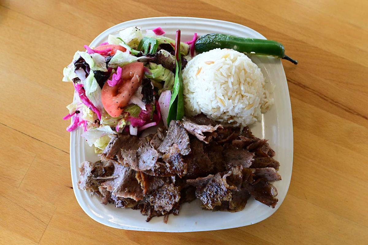 A plate with sliced grilled meat, a mound of white rice, a fresh salad with lettuce, tomato and onions, and two green chili peppers, served on a wooden table.