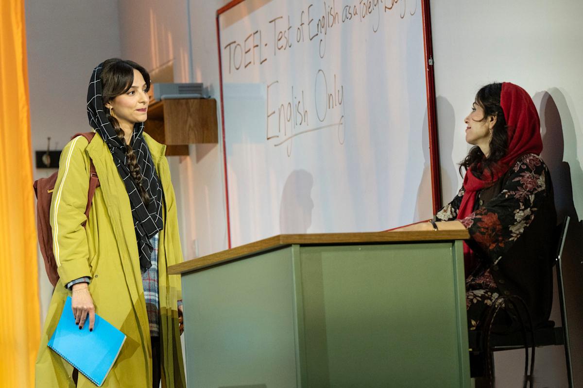 Two women talk in a classroom. One stands holding a blue folder, wearing a yellow coat and headscarf. The other sits at a desk, wearing a patterned dress and red scarf. A board behind them reads “TOEFL: Test of English.”.