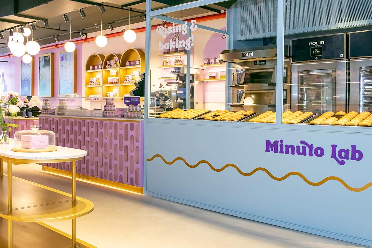 A modern bakery interior with pastel-colored decor, a display of freshly baked bread behind glass, and a counter with pink and purple accents. The sign reads "Minuto Lab" and a cake is on a table in the foreground.