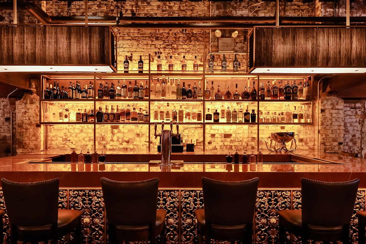 A stylish bar with ornate chairs, a polished counter, and shelves of assorted liquor bottles against an exposed brick wall, warmly lit with golden lighting.