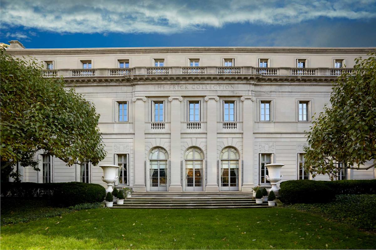 The image shows the facade of The Frick Collection, a grand white neoclassical building with tall windows and ornate details. It is flanked by neatly trimmed bushes and trees under a partly cloudy blue sky.
