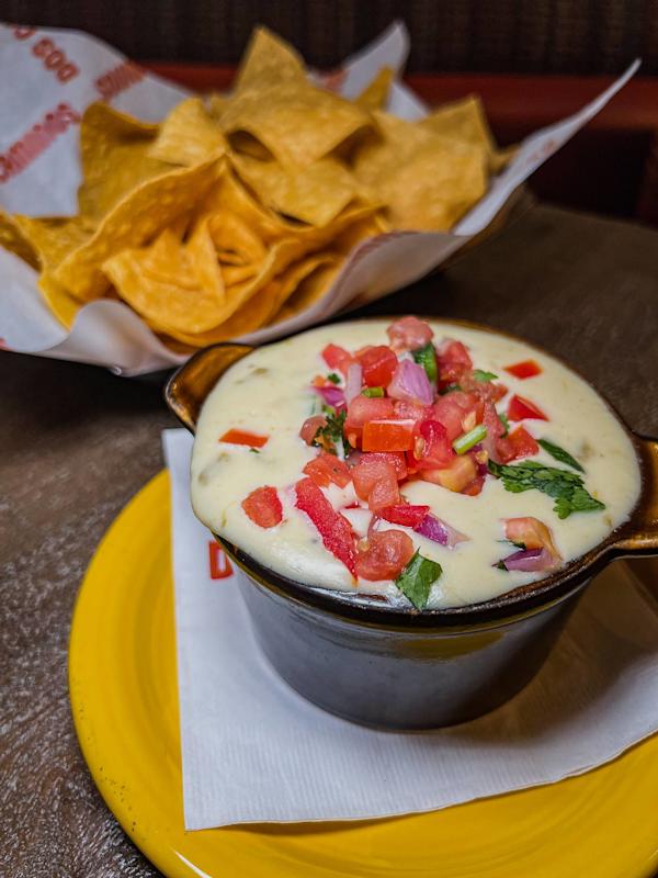 A bowl of creamy queso dip topped with diced tomatoes, onions, and cilantro sits on a yellow plate, with a basket of tortilla chips in the background.