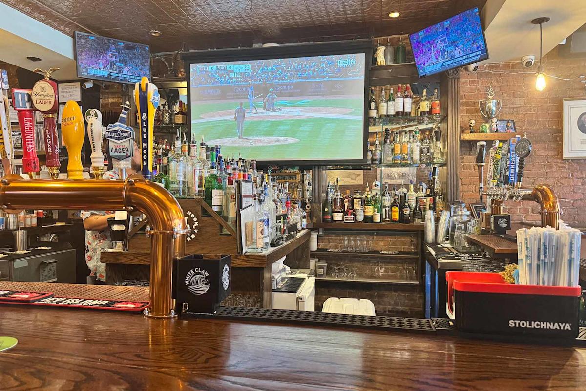 A bar with various beer taps, liquor bottles, and drink supplies on the counter. A large TV above the bar shows a baseball game, and two smaller TVs display sports. The bar has a cozy, casual atmosphere.