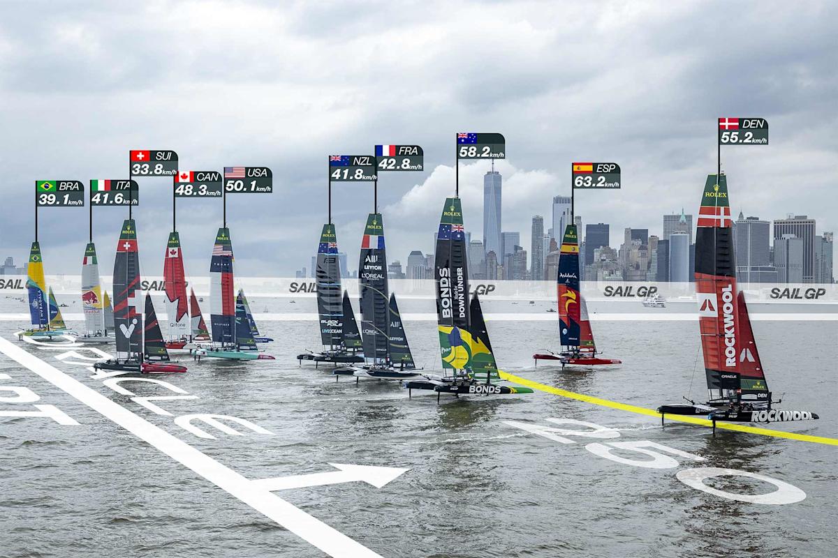 A group of high-tech sailboats race on the water near a city skyline, each boat displaying country flags and speed data above them. The image shows a competitive sailing event with various nations participating.