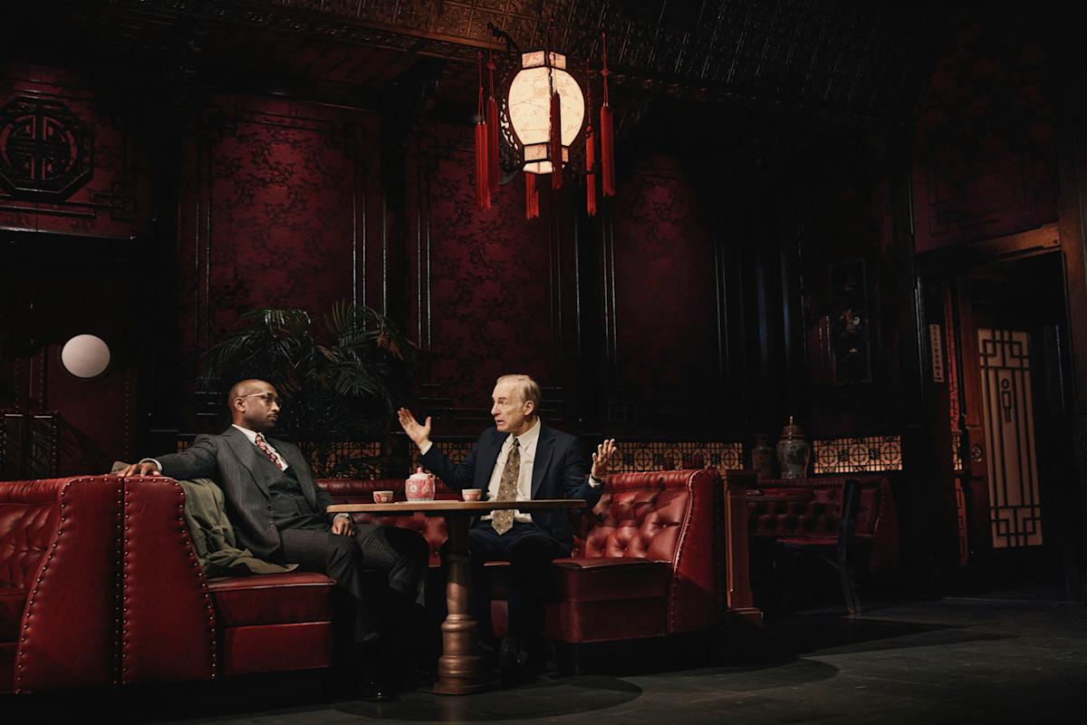 Two men in suits sit in a dimly lit, ornately decorated restaurant booth with red leather seats and dark red walls, having a serious conversation under an elaborate hanging lantern.
