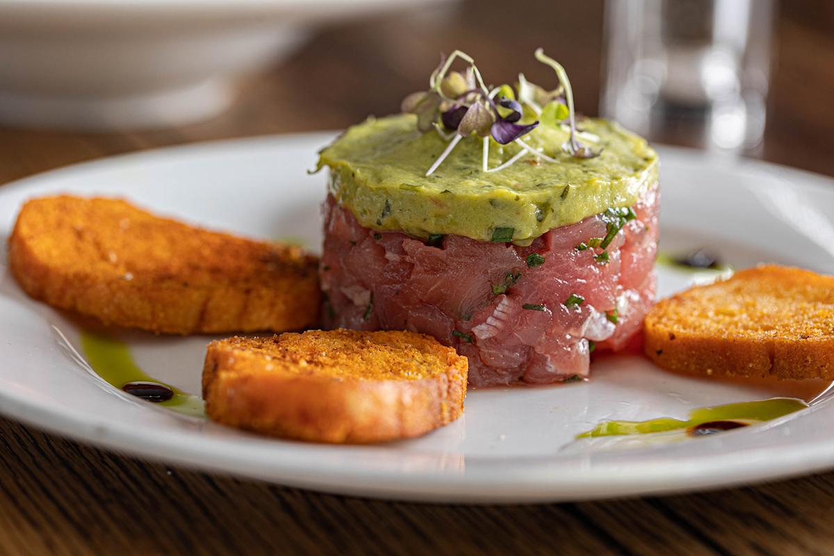 A plate with tuna tartare topped with avocado and microgreens, surrounded by three toasted bread slices and decorative drizzles of sauce.