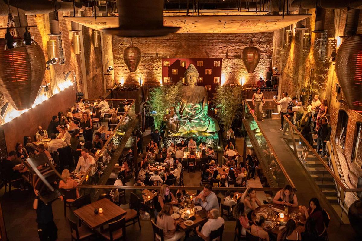 A lively restaurant with many diners seated at tables on two levels, warm ambient lighting, exposed brick walls, and a large golden Buddha statue as the central focal point.