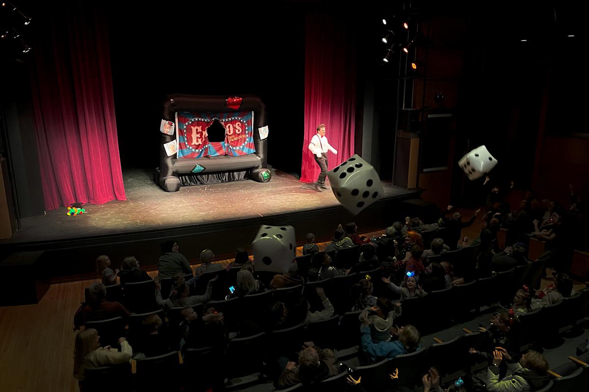 A performer stands on a stage in front of a red curtain, engaging an audience. Large inflatable dice are being tossed around in the crowd. The stage features a colorful backdrop labeled "Eros." The auditorium is filled with seated spectators.