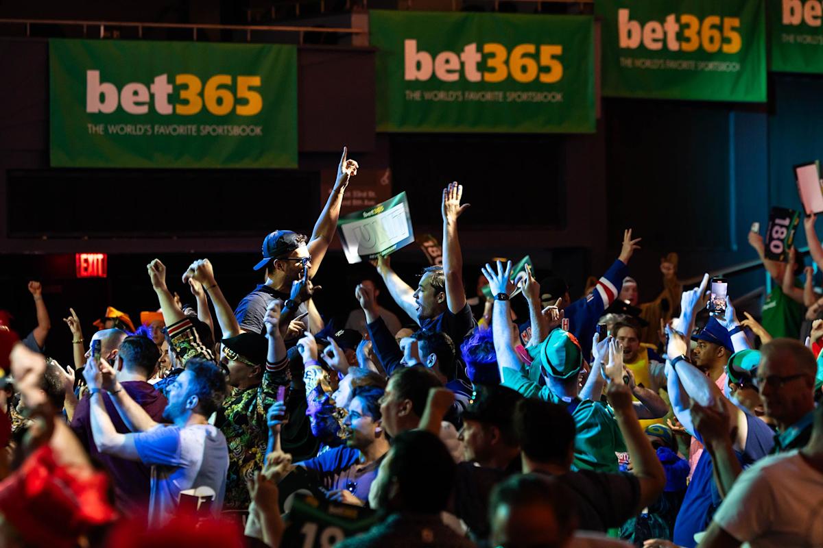 A large, energetic crowd cheers and raises their arms at an indoor sports event, with prominent green “bet365” banners hanging in the background. The atmosphere is festive and lively.