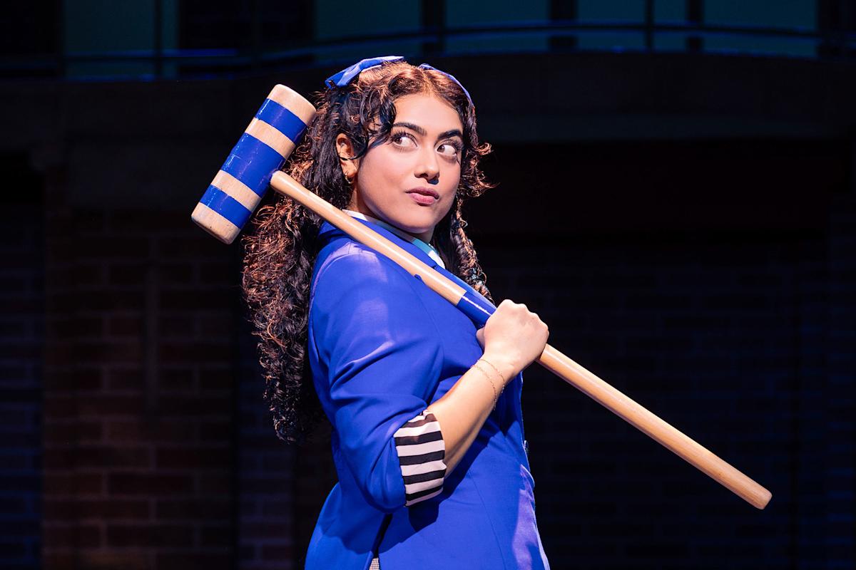 A woman with long curly hair, dressed in a bright blue outfit, holds a large wooden mallet with blue stripes over her shoulder and looks to the side on a stage with a dark background.