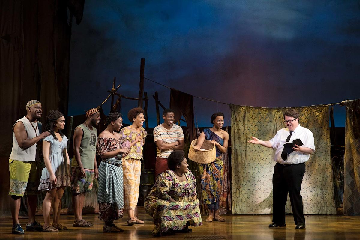 A group of people in colorful clothing stand on a stage, listening to a man in glasses holding a book and gesturing. The background suggests an outdoor village scene with draped fabric and props.