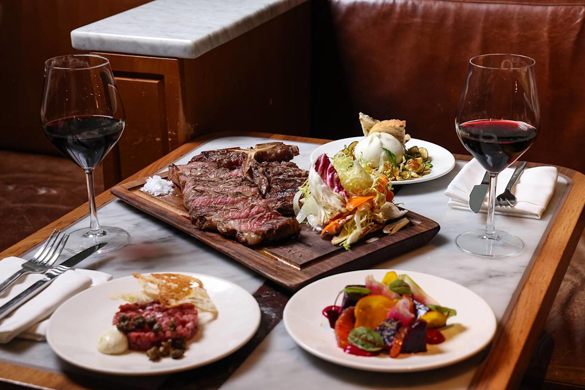 A table set for two with glasses of red wine, a wooden board with grilled steak and salt, and plates of salad, vegetables, and a meat appetizer in a cozy restaurant booth.