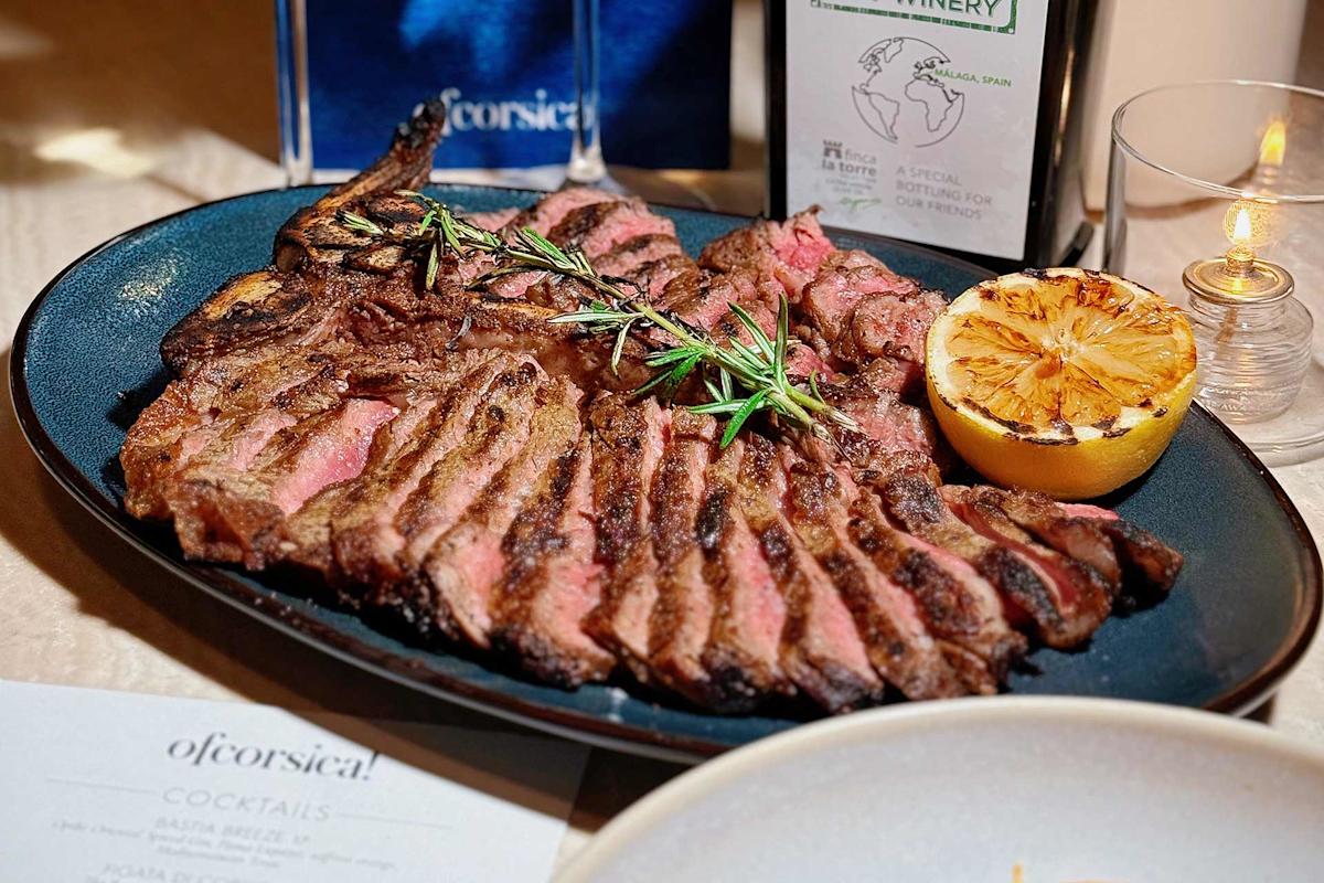 A sliced grilled steak garnished with rosemary is served on a dark oval plate with a charred lemon half. A cocktail menu, wine bottle, and candle are visible in the background.