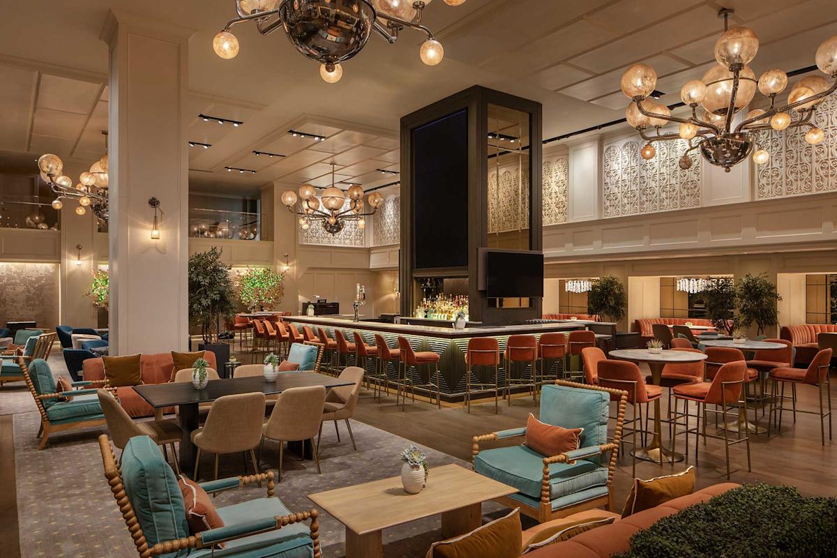 Spacious, elegant hotel lounge with modern chandeliers, a central bar surrounded by orange stools, and various seating areas featuring teal, orange, and beige chairs, wooden tables, and potted plants throughout.