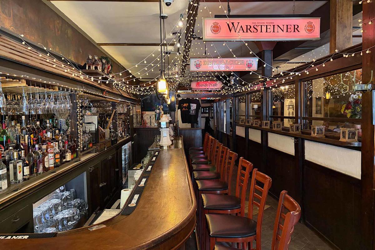 A cozy bar with wooden chairs lined along a curved counter, shelves stocked with bottles and glasses, string lights hanging from the ceiling, and Warsteiner signs above the bar. The atmosphere is warm and inviting.