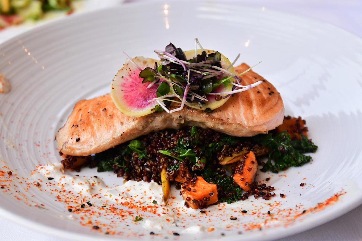 Grilled salmon fillet served atop a bed of quinoa and sautéed vegetables, garnished with microgreens and a thin radish slice on a white plate, with a sprinkle of seasoning around the edges.