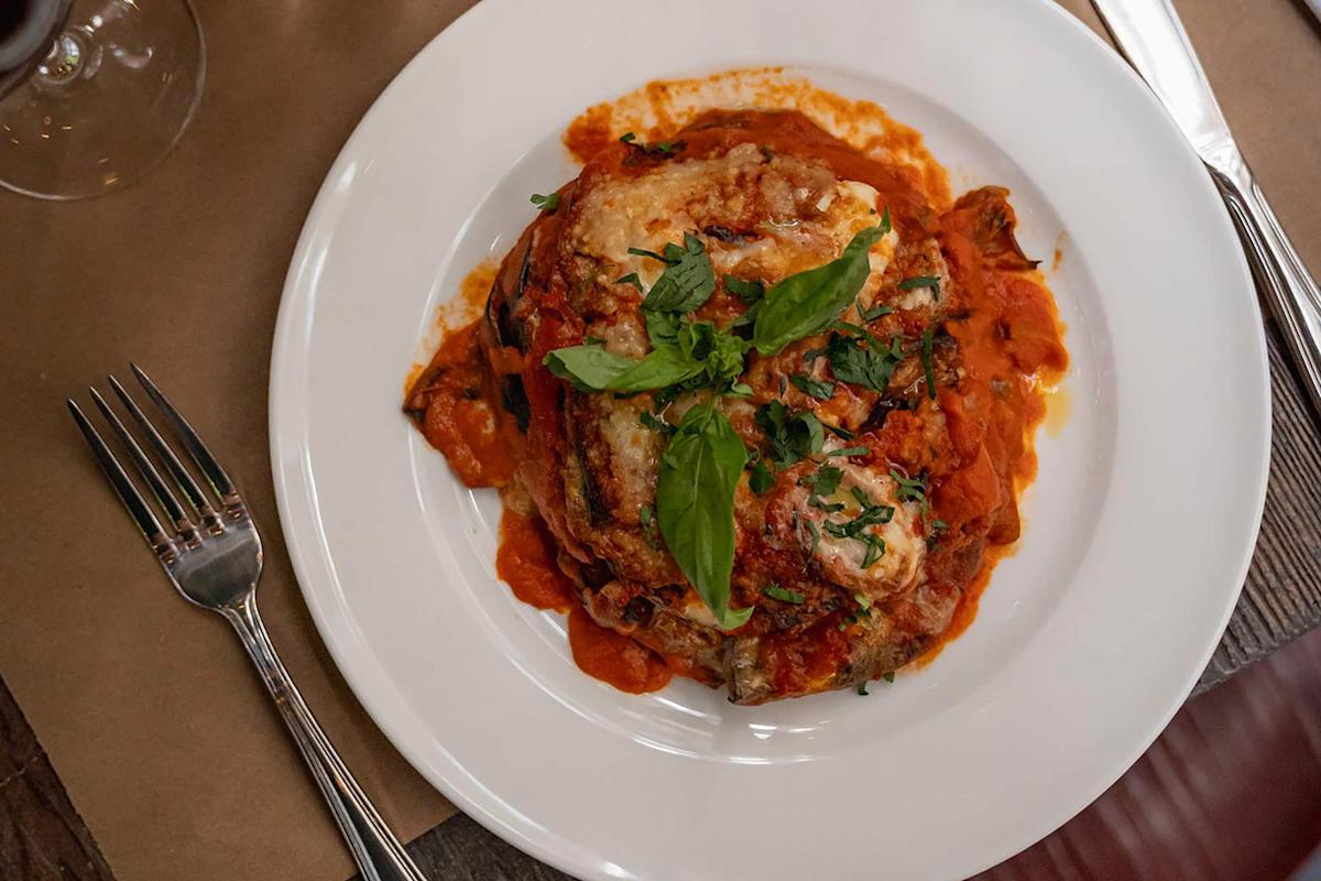 A white plate with eggplant parmesan topped with tomato sauce, melted cheese, and fresh basil, set on a brown placemat with a fork and knife beside it.