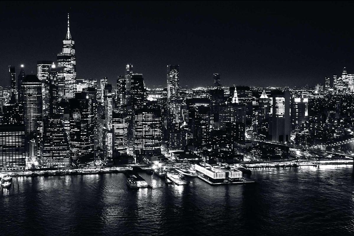 Black and white aerial view of the Manhattan skyline at night, featuring tall skyscrapers with illuminated windows reflecting on the water below.
