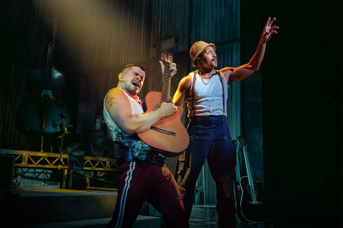 Two men perform on stage; one passionately plays an acoustic guitar, while the other raises his arm dramatically. Both wear casual clothes and are lit by stage lights, with musical instruments visible in the background.