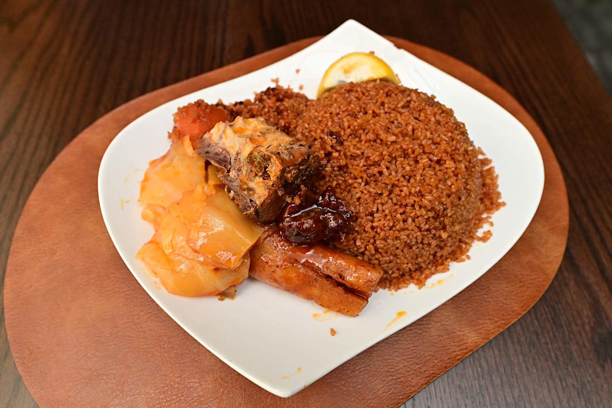 A plate of jollof rice with a serving of stewed meat, vegetables, and a slice of lemon, presented on a white dish atop a wooden table.