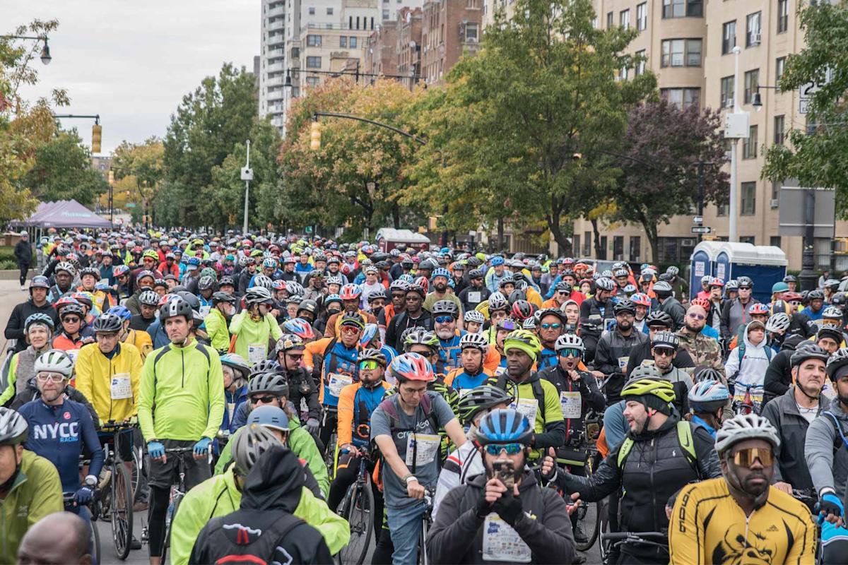 A large crowd of cyclists wearing helmets and colorful gear gathers on a city street lined with trees and tall buildings, preparing for a group ride or cycling event.